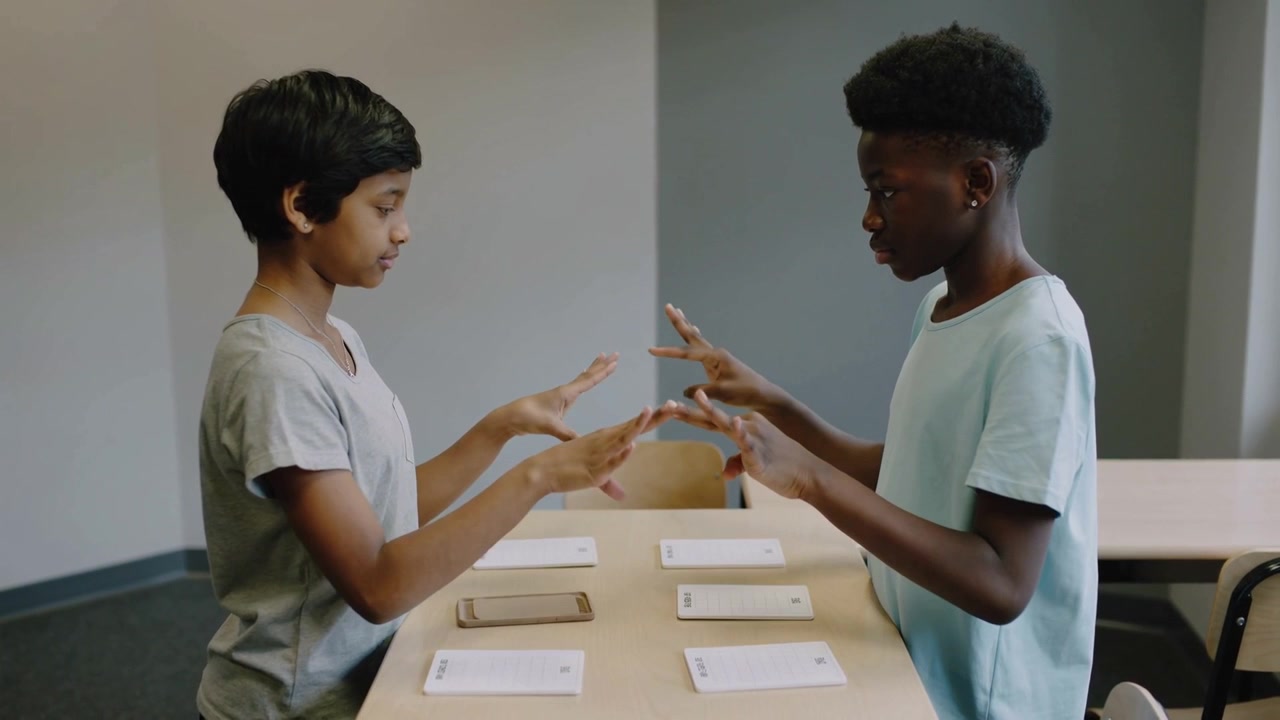 Picking up token left grey-tee teen practicing passing to right blue-tee at school table cards cell