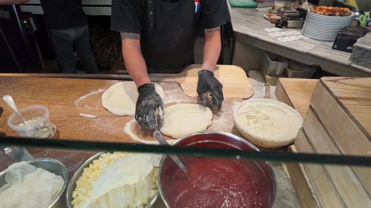 Uno chef con guanti neri stende l'impasto fresco della pizza su un bancone da cucina in legno infarinato.