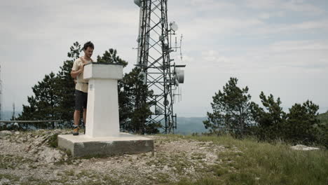 excursionista con una mochila naranja parada junto a un monumento de hormigón en la cima de la montaña slavnik cerca de la torre de radio