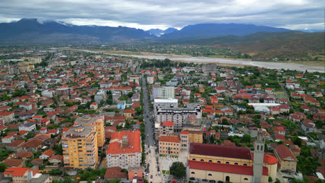 aerial drone forward moving shot over city buildings with river flowing in the background along mountain range on a cloudy day