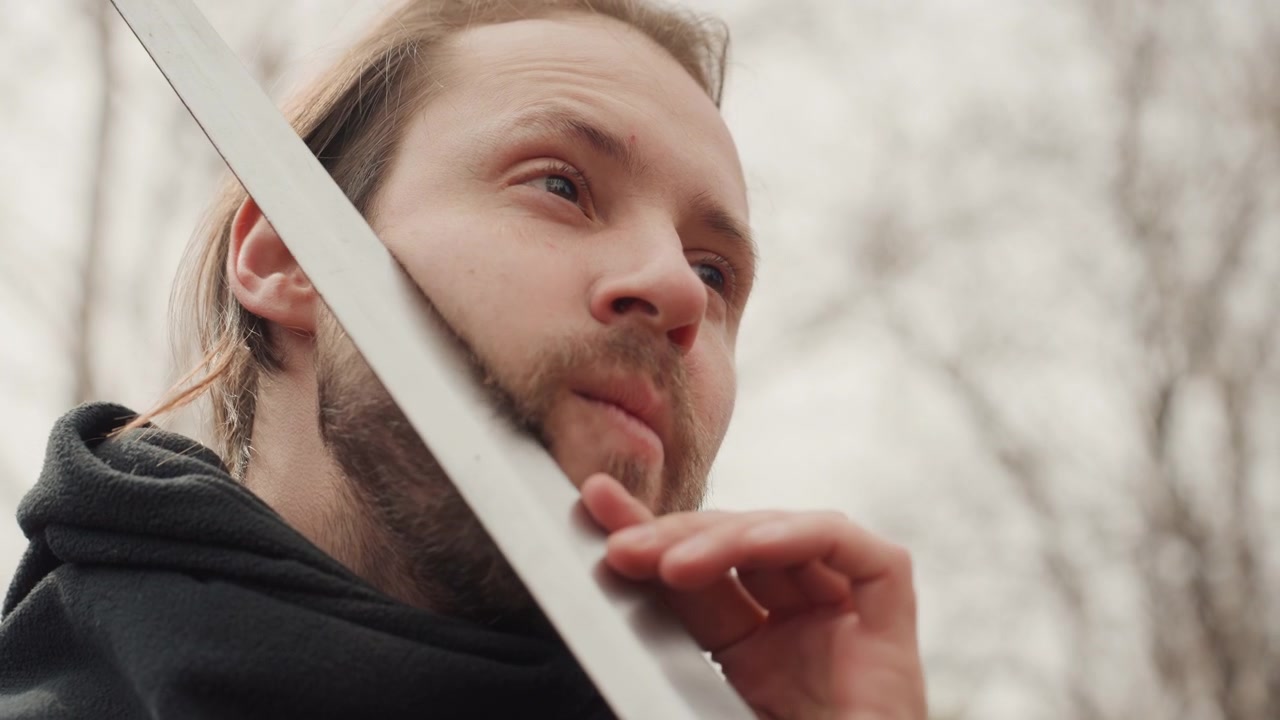 Adult man outdoors holds metal sword across chest near face while inspecting blade and hilt, using careful hand gestures, slight head tilts, and focused gaze to show calm readiness and concentration