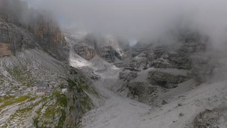 volando a través de nubes grises que revelan montañas rocosas y cabañas de excursionistas en los dolomitas