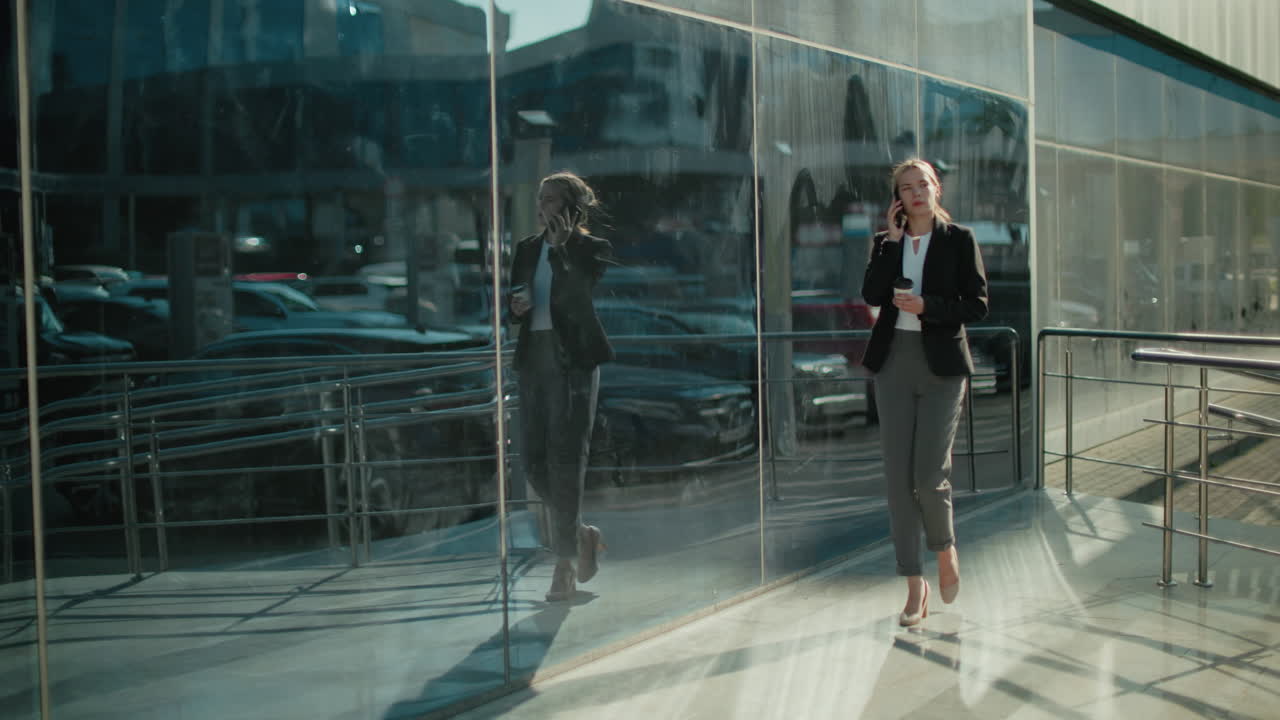 Elegant lady walking confidently with coffee in hand while on phone call, reflected on large glass building under bright sunlight, showcasing urban lifestyle and professional morning routine