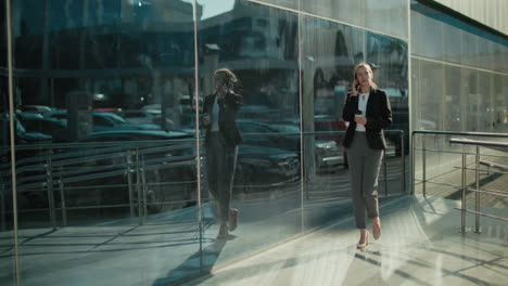 elegant lady walking confidently with coffee in hand while on phone call, reflected on large glass building under bright sunlight, showcasing urban lifestyle and professional morning routine