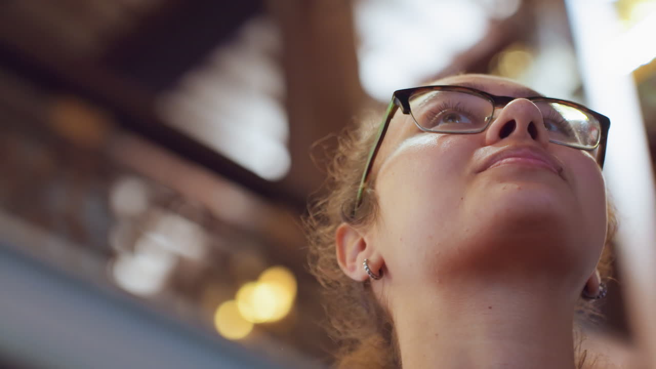 Mujer latina mirando hacia el techo, la escena captura la mirada ascendente, vigas y arcos arquitectónicos, cálida luz de linterna, silueta con capucha, ambiente de viajera curiosa, expresión reflexiva, museo o centro comercial.