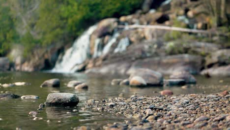 Felsiges-Flussufer-Mit-Sanften-Wellen,-Die-über-Die-Küste-Strömen,-Mit-Glatten-Steinen-Und-Fließendem-Wasserfall-Im-Hintergrund