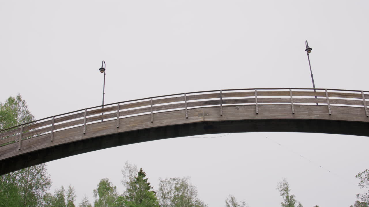 Low-angle view of arched wooden pedestrian bridge spanning river surrounded by tall leafy trees, captured from water surface during overcast day in quiet Norwegian wilderness