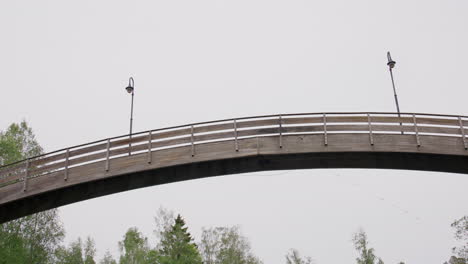 low-angle view of arched wooden pedestrian bridge spanning river surrounded by tall leafy trees, captured from water surface during overcast day in quiet norwegian wilderness