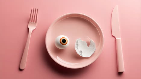 surreal eyeball and eggshell on pink plate with cutlery