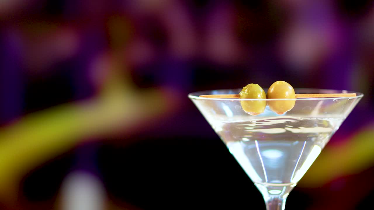 A martini glass with olives against a colorful, blurred background. Bright lighting highlights the drink's clarity and elegance