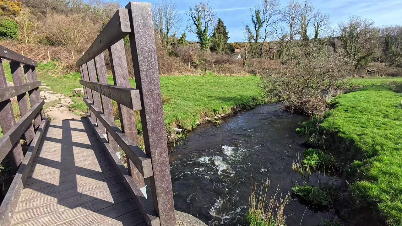 Sunlit wooden bridge crossing over flowing clean stream in welsh countryside meadow field