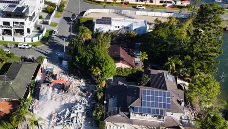 drone footage captures a demolition excavator tearing down a residential house in a suburban gold coast neighborhood, with debris and dust visible in bright daylight