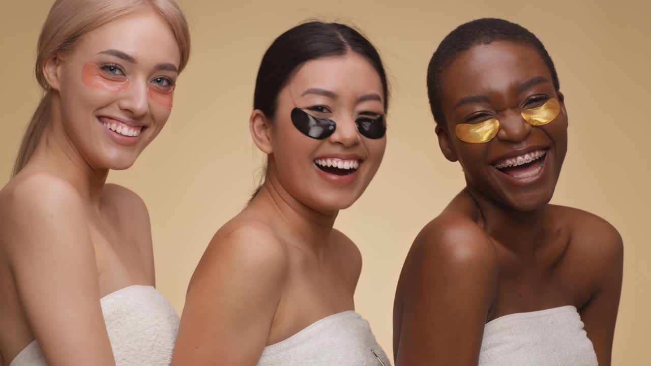 Portrait en gros plan de trois jeunes femmes diverses avec des patchs oculaires au collagène, riant à la caméra, posant enveloppées dans des serviettes