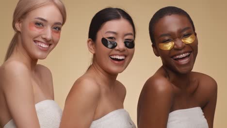 close up portrait of three young diverse ladies with collagen eye patches laughing to camera, posing wrapped in towels