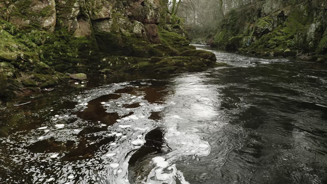 Swirling bubbles of white foam float slowly on the surface a Scottish river in a hypnotic, constantly changing pattern of organic shapes as a fast river current flows through a green, mossy gorge
