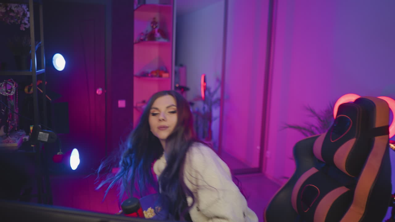 Gamer walks into studio carrying bowl of popcorn heading toward ergonomic chair surrounded by neon lights and gaming gear, preparing to sit and adjust position for cozy entertainment session