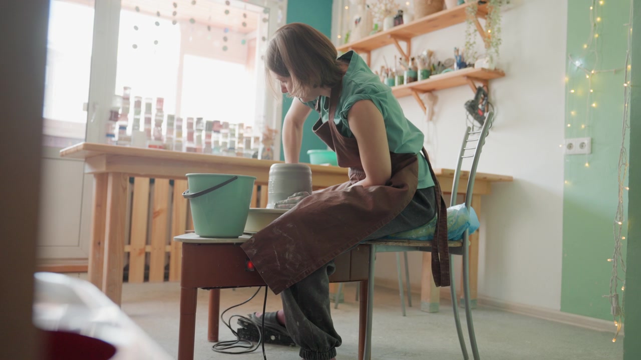 White woman shaping clay on wheel in sunlit pottery workshop wearing apron bucket beside jars on shelf fairy lights soft green walls focused hands molding vessel intimate studio atmosphere