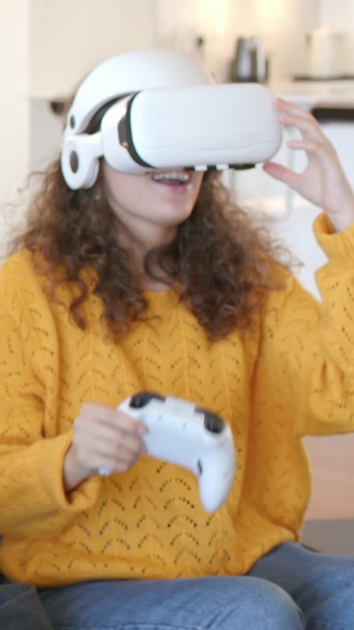 Donna con maglione giallo che reagisce a contenuti VR, impugnando un controller sul divano e regolando il visore.