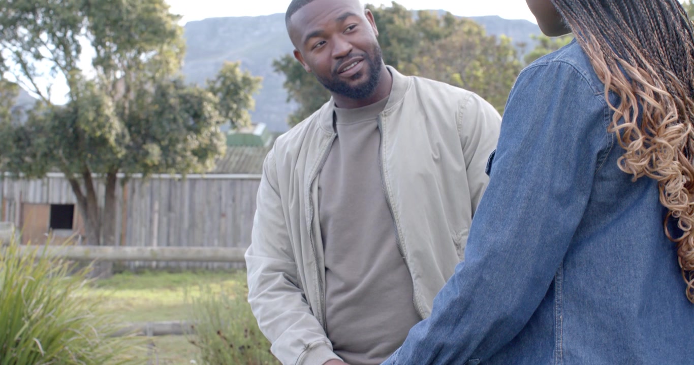African couple in denim, tan jackets holding green cloth, adjusting, man shifting, chatting in yard