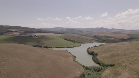 Drohnenaufnahme-Von-Kurvenreichen-Straßen-Und-Goldenen-Feldern-In-Der-Landschaft-Der-Toskana,-Italien,-An-Einem-Sonnigen-Tag-Mit-Blauem-Himmel-Und-Wolken-Am-Horizont-Und-Einem-Kleinen-See-Oder-Einer-Pfütze-In-Der-Mitte-Des-Szenenprotokolls