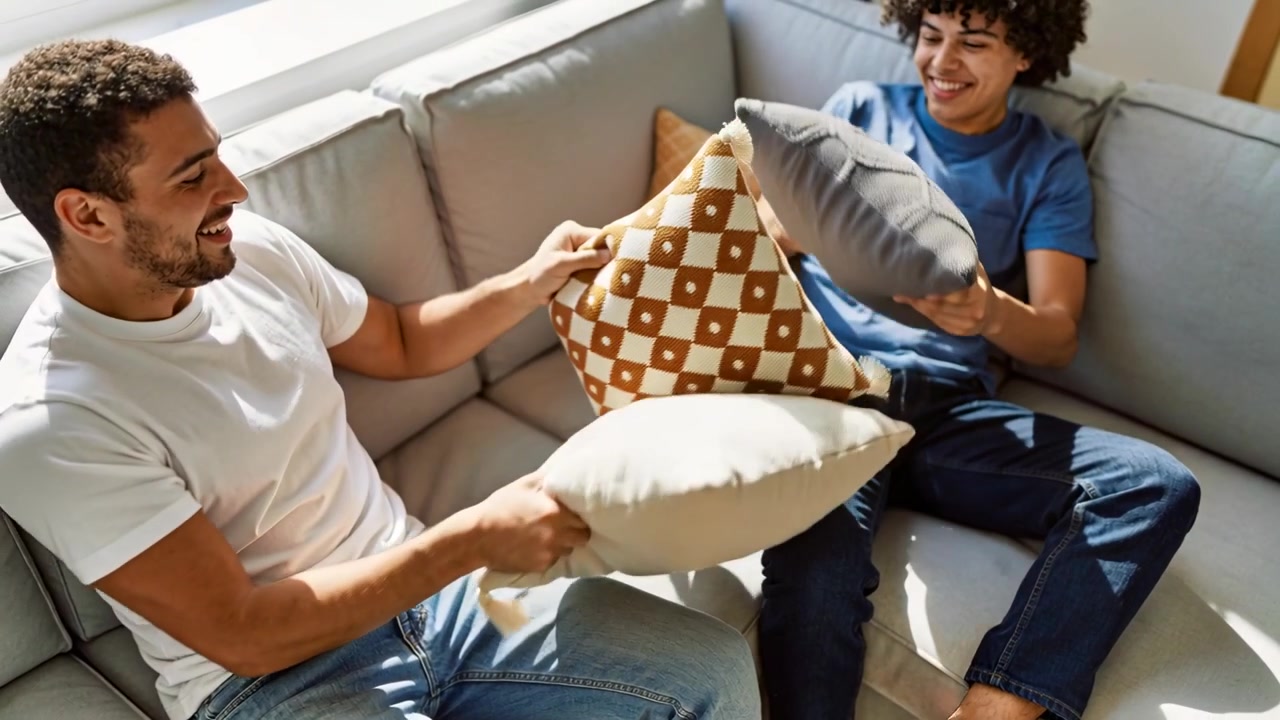 Papà e figlio con magliette bianche e blu che sollevano cuscini, iniziando una battaglia di cuscini su un divano componibile.