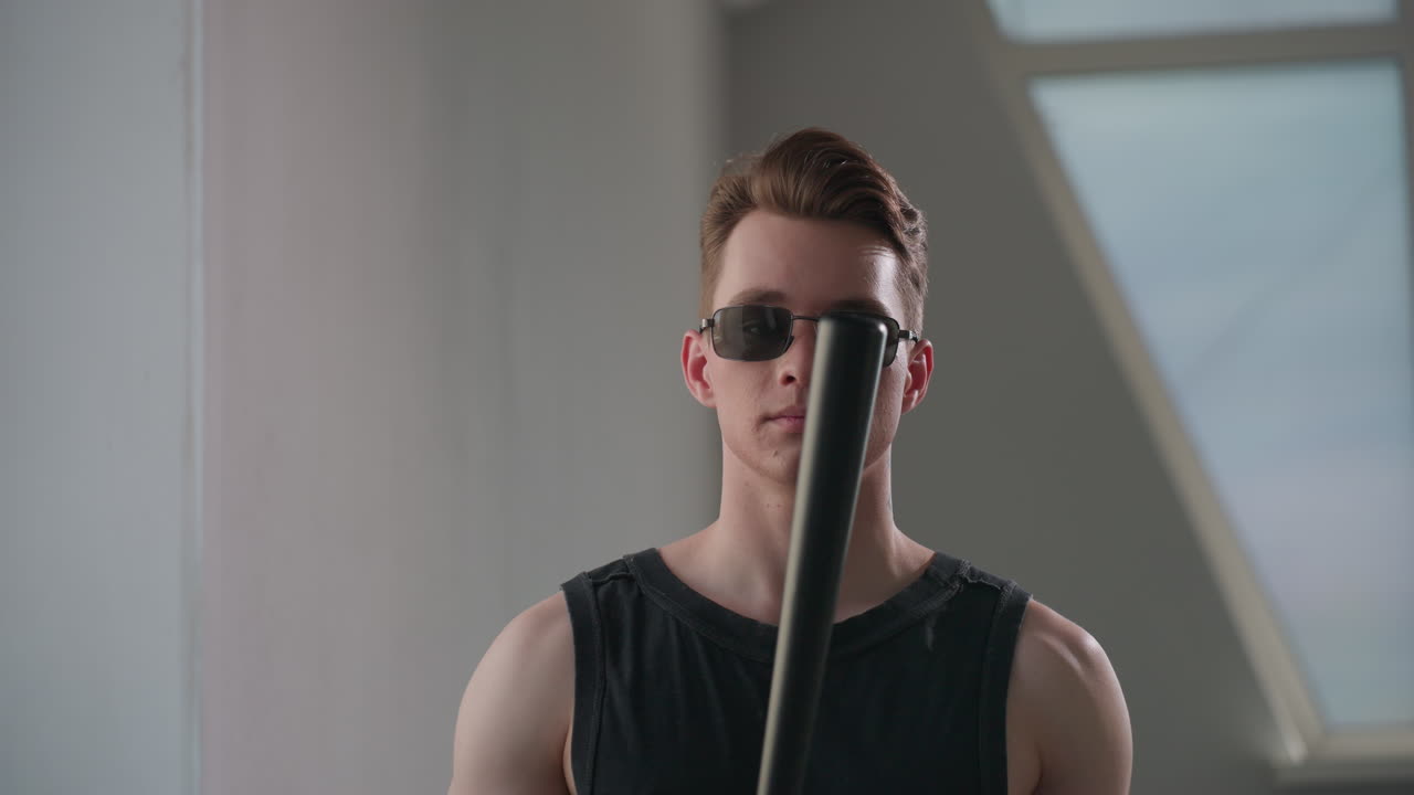 Boy in black tank top and dark sunglasses examines metal security baton by loft window, light filtering through panes onto tattooed neck and toned arms, conveying modern style