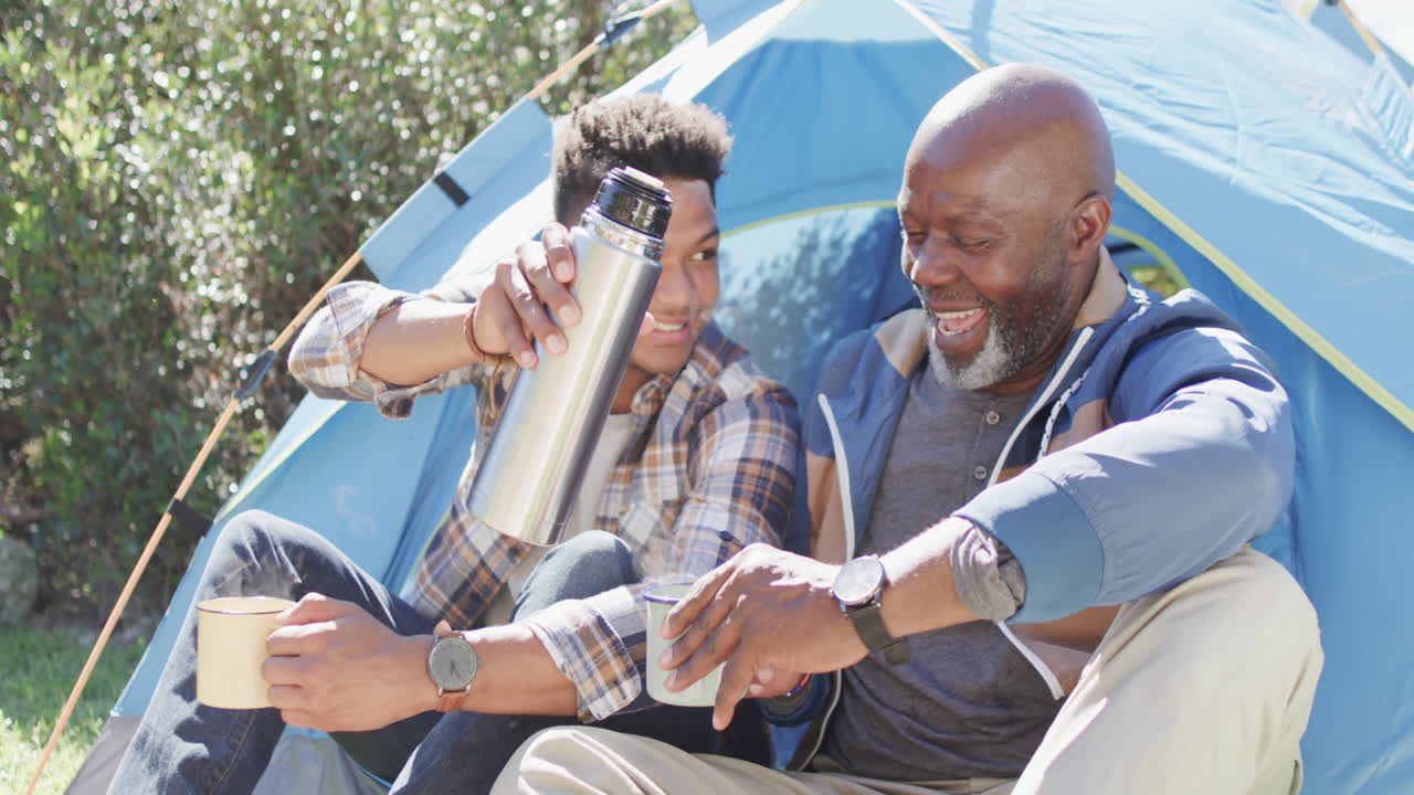 Happy african american father and adult son sitting outside tent drinking coffee in sun, slow motion