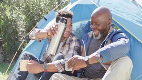 happy african american father and adult son sitting outside tent drinking coffee in sun, slow motion