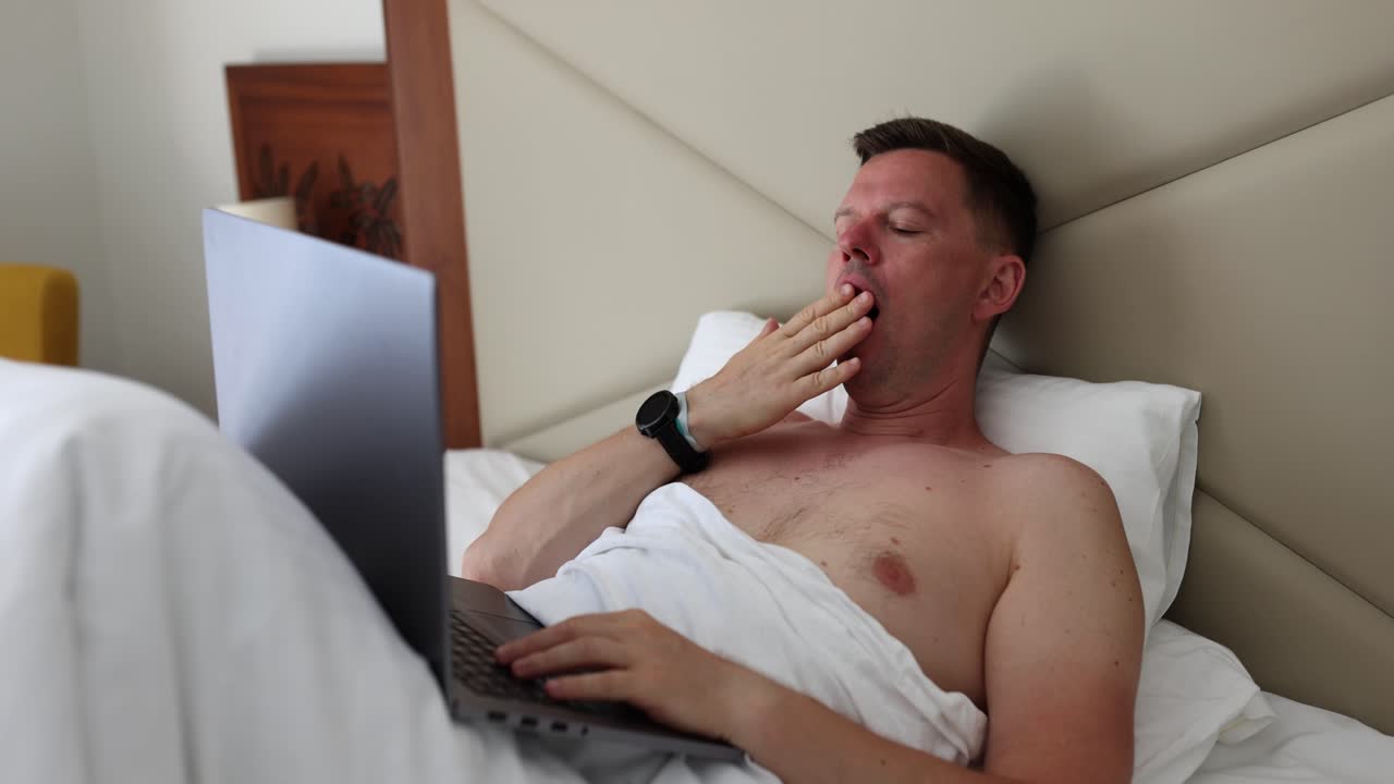 Tired man yawning in bed while using a laptop
