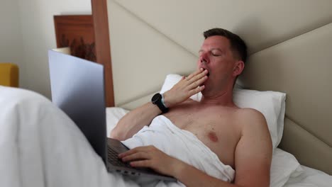 tired man yawning in bed while using a laptop