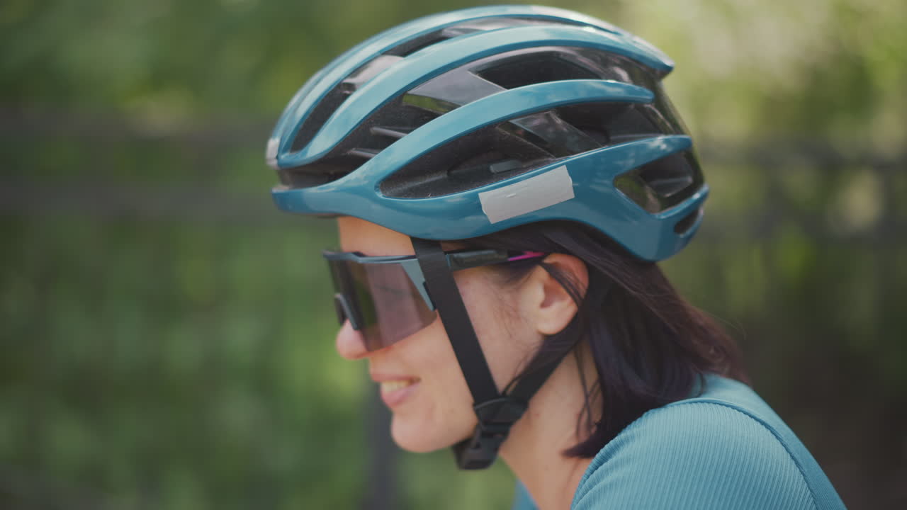 Woman cyclist wearing teal helmet and sunglasses, side profile in park smiling portrait with short dark hair tucked under helmet, chin strap fastened, reflective visor, relaxed solo ride