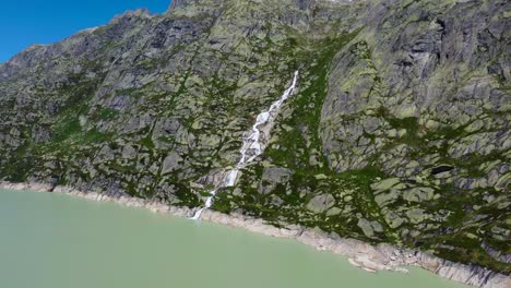 Luftdrohnenschwenkaufnahme-über-Einem-Wasserfall-In-Der-Ferne-Entlang-Des-Grimselsees-In-Der-Nähe-Der-Grimselpass-Hochgebirgsstraße-In-Den-Wunderschönen-Schweizer-Alpen,-Schweiz-Bei-Tag