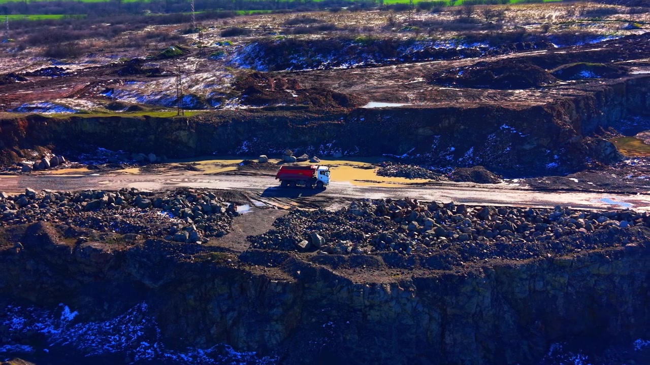 Wide aerial view of rock quarry pit with dump truck, Bulgaria. Layered rock walls, muddy pond and green fields beyond the pit edge.