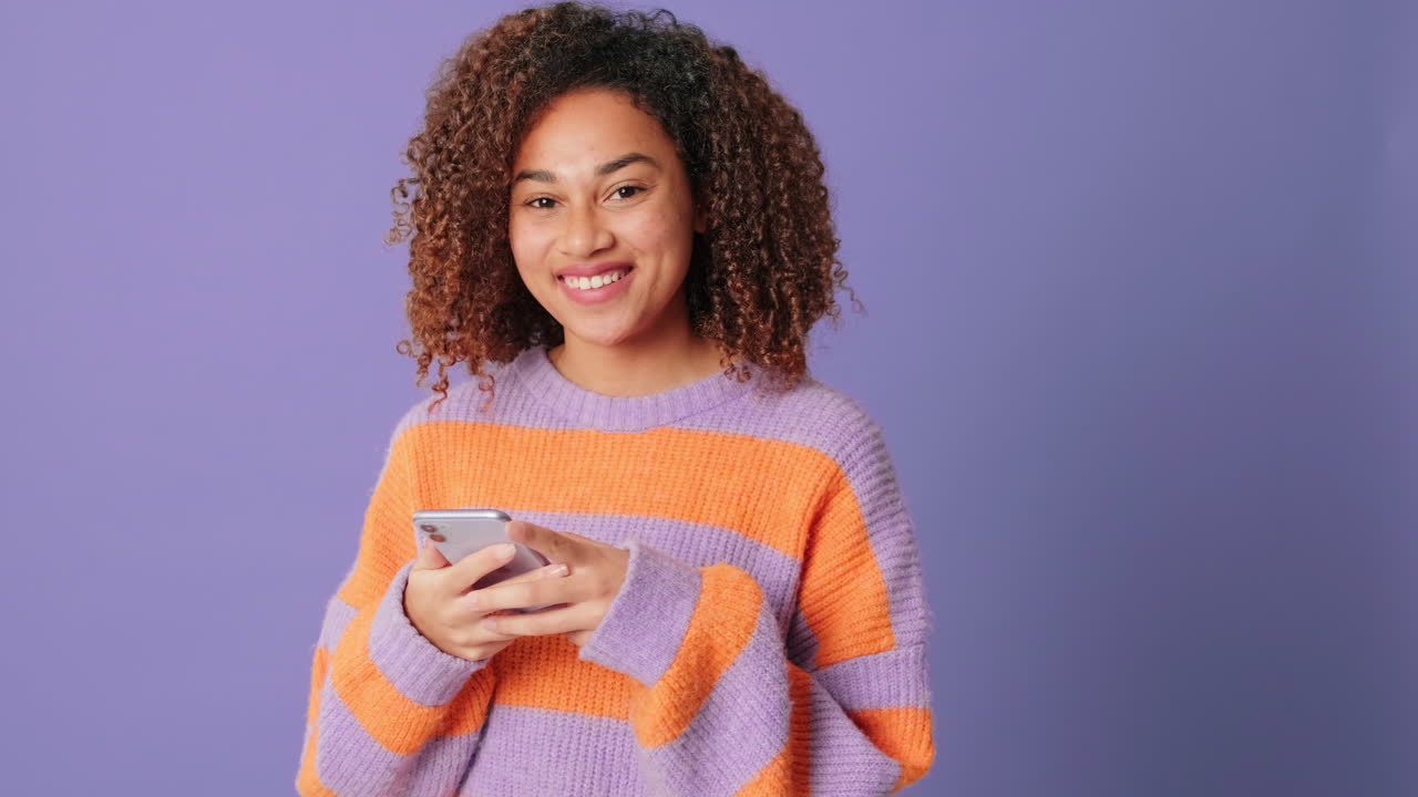 Smilende ung kvinde bruger smartphone på lilla baggrund