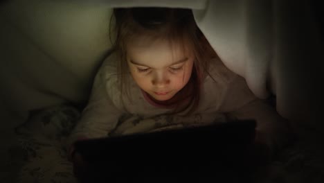 niño leyendo en la tableta bajo la manta por la noche