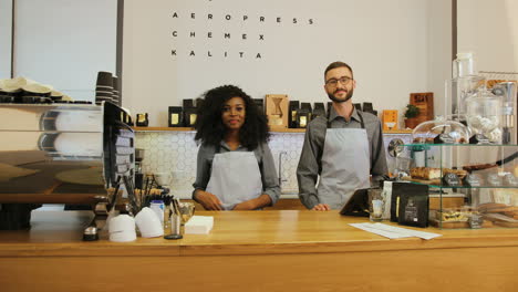 young caucasian barista and her african american coworker posing for the camera and crossing arms behind the bar of a coffee shop