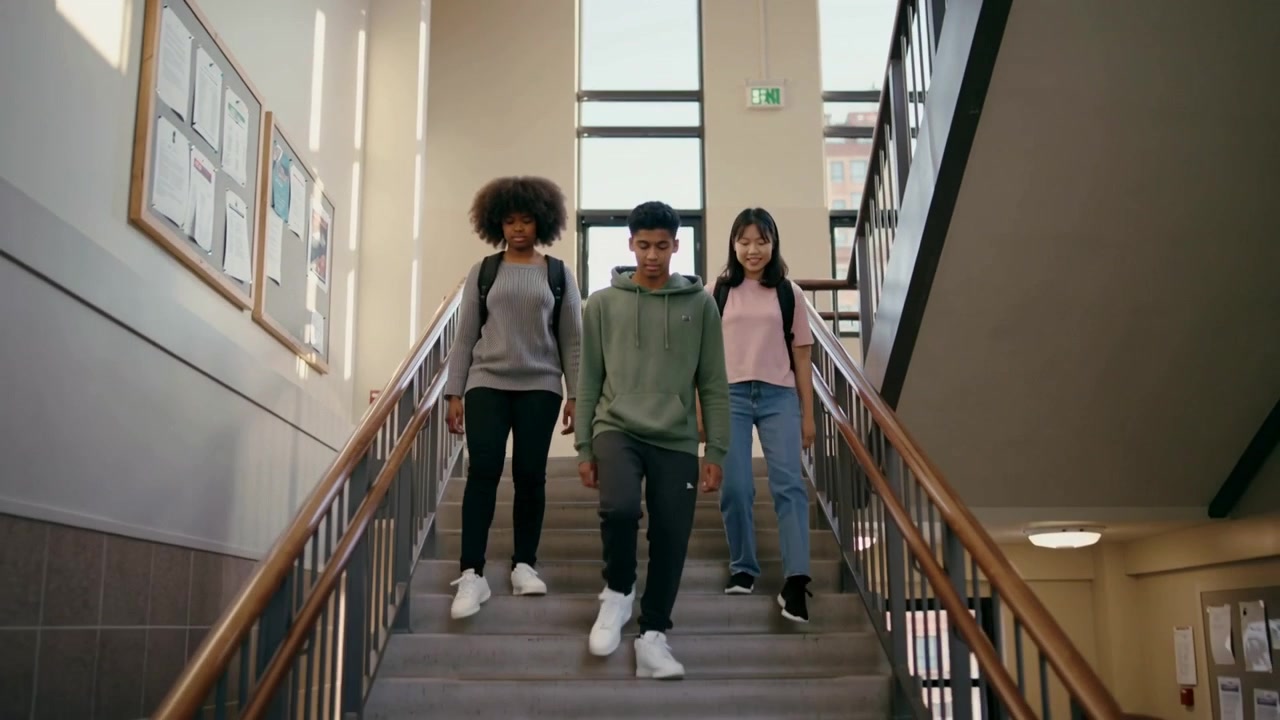 Tre studenti in abiti casual scendono le scale della scuola, portando gli zaini e passando davanti alle finestre.