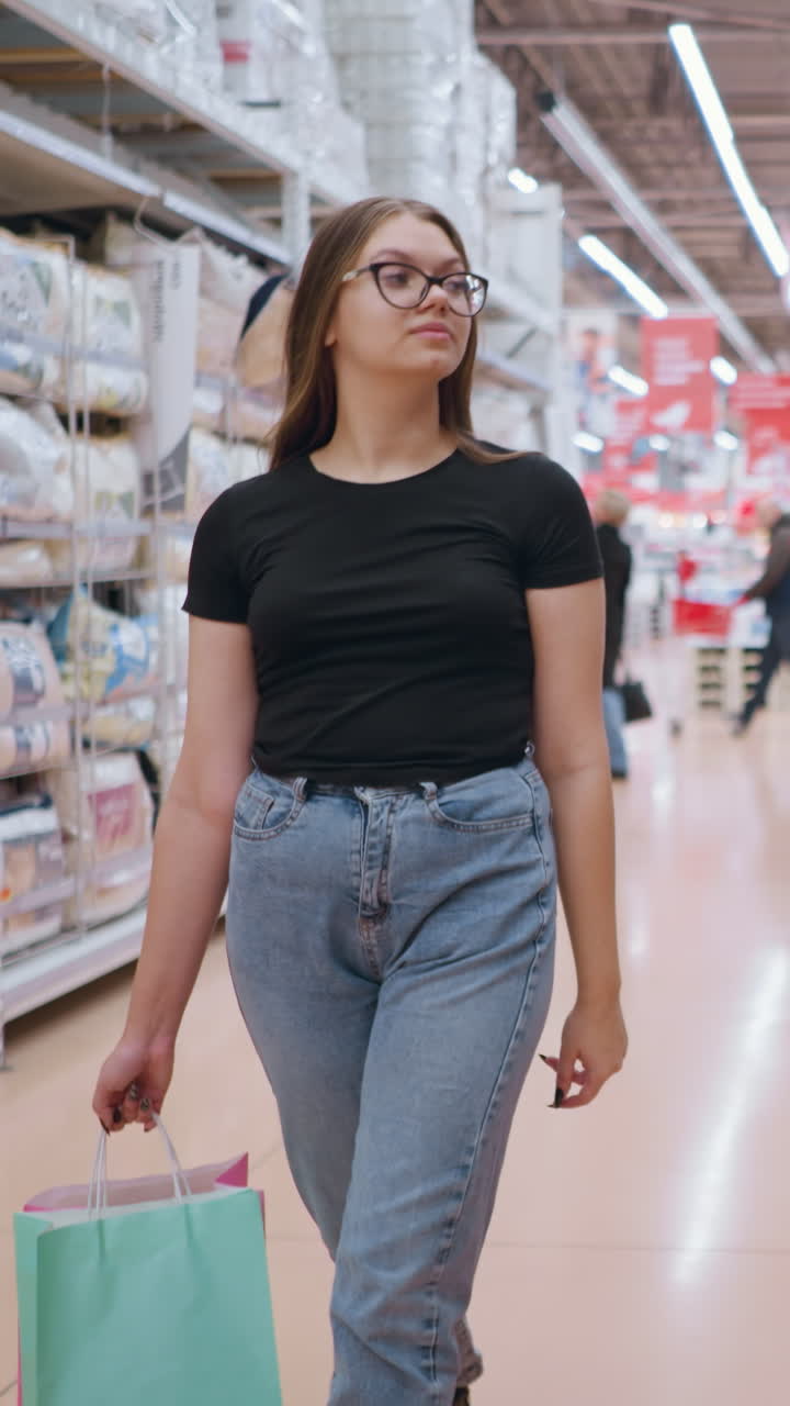 검은 과 청바지를 입은 여성이 섬유 제품 근처의 슈퍼마켓 통로를 걸어 가며, 쇼핑 가방을 들고 선반에 전시된 상품에 초점을 맞추고, 밝은 소매 환경