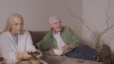 happy senior woman playing video game sitting on sofa at home while enthusiastic elderly man supporting her