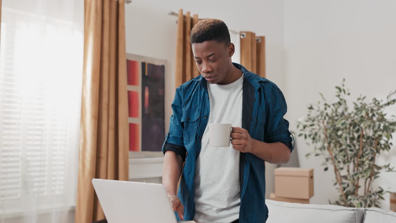 Buying furniture on the Internet, happy smiling man drinking coffee and clicking on a laptop standing on cardboard boxes, a boy has moved into a new apartment and is wondering how to furnish the house