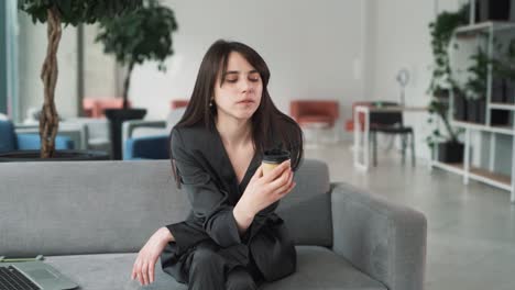 young and beautiful woman in a suit sits on the couch in the business center, pondering silently while looking at a cup of coffee