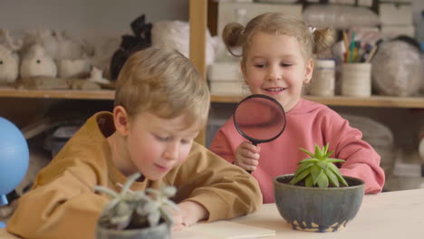 petite fille observant une plante avec une loupe assise à table près d'un ami qui dessine dans un atelier d'artisanat 1