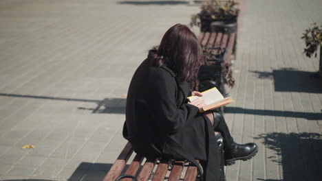 lady in black coat and leather boots sits on bench in public, leg crossed, reading book, flower pots in background. urban street with plants and clear sky in the scene