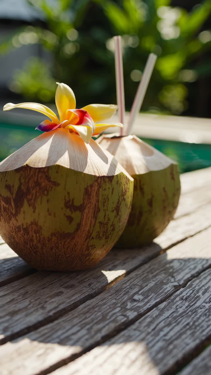 Close-up, eye-level shot of two coconuts with straws and a flower, set on a wooden table by a pool