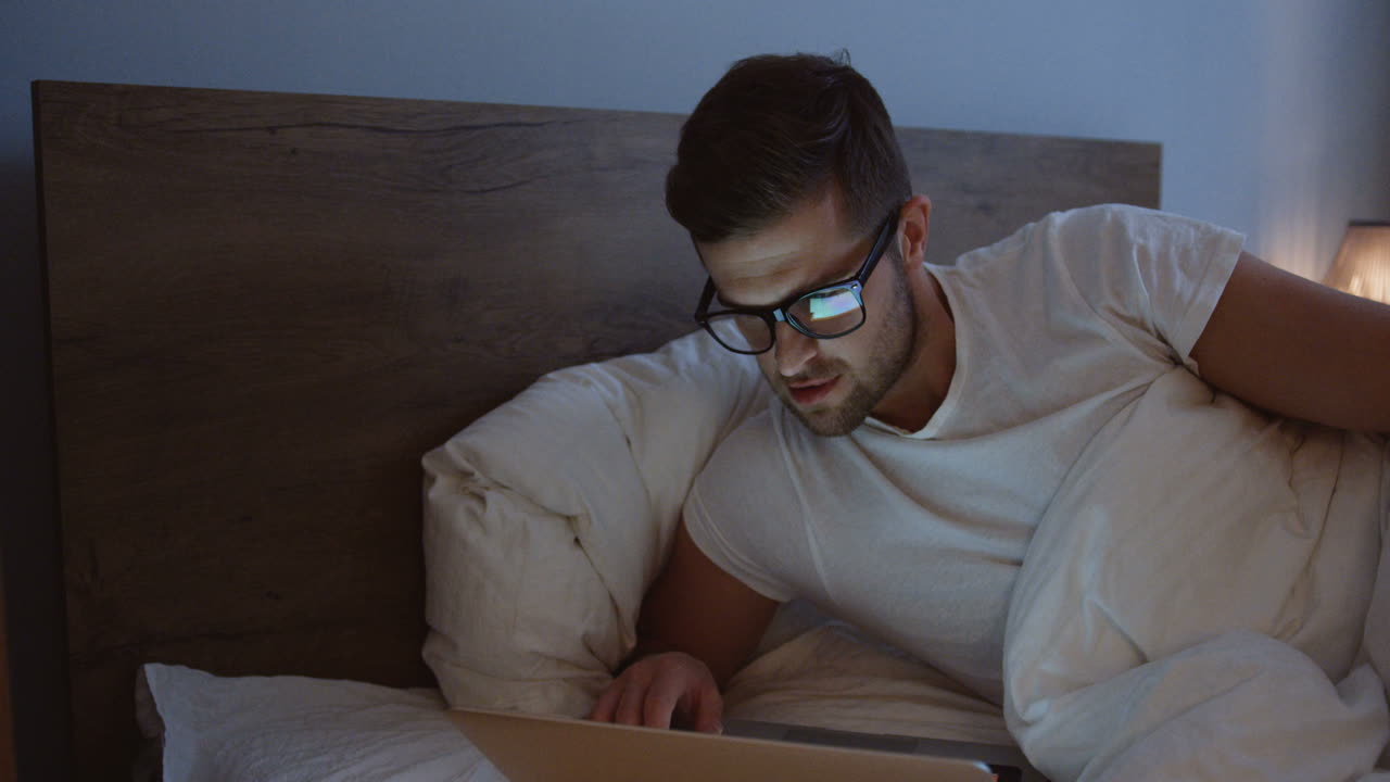 Close-up view of smiling man using laptop and smiling while watching something in the evening in her bed