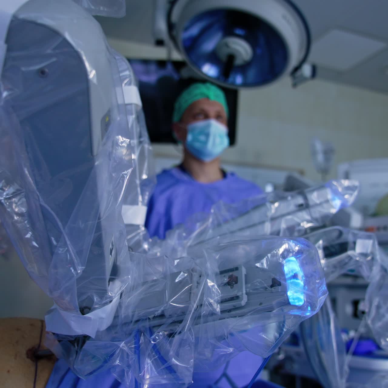Manipulator arms of Da Vinci surgeon system coated in plastic move slowly at procedure. Male doctor in mask stands behind the machine at backdrop in blur