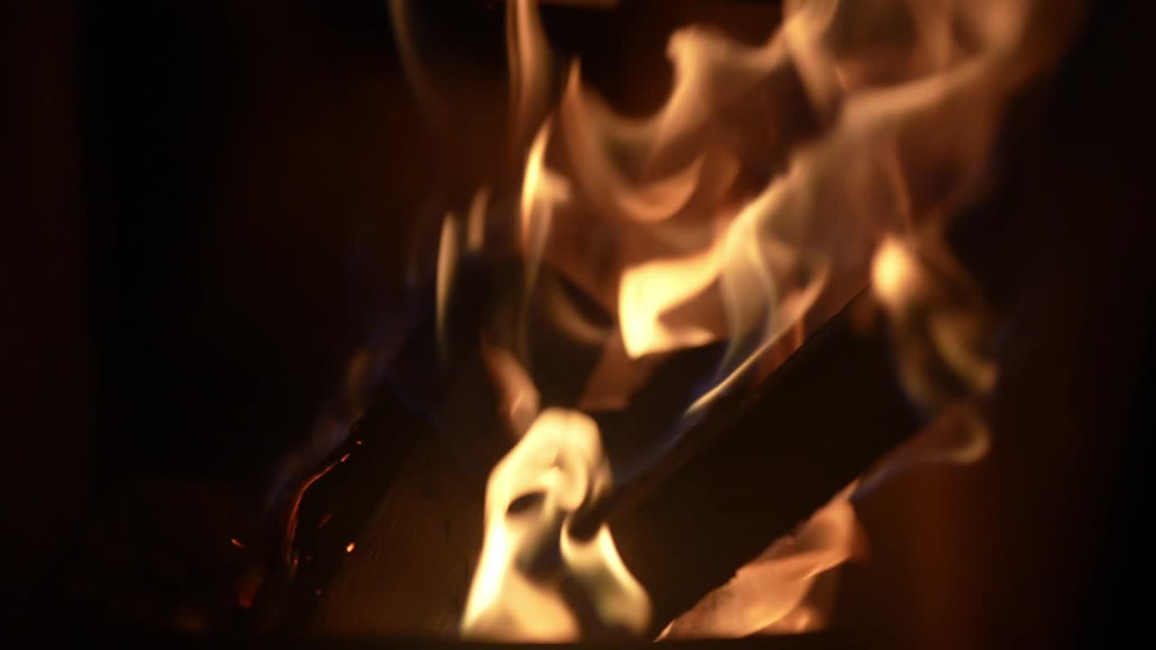 A campfire burns in the night, flames dancing slowly, casting flickering shadows. Sparks rise into the darkness, the warmth contrasting with the cold air.