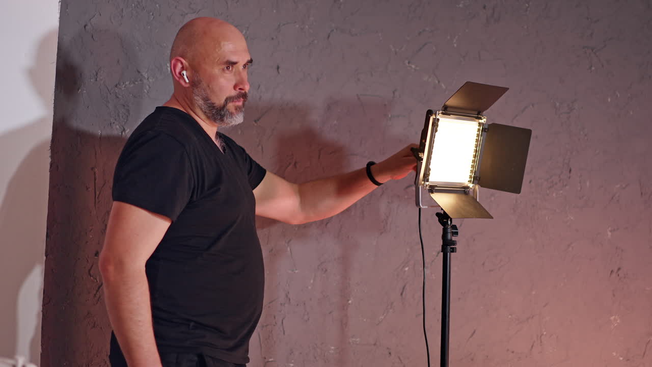 Bald man with grey beard stands near the soffit. Cameraman setting the light equipment in studio.