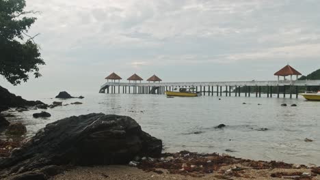 playa rocosa con barco pasando y embarcadero en el fondo durante el día nublado y soleado en malasia, asia
