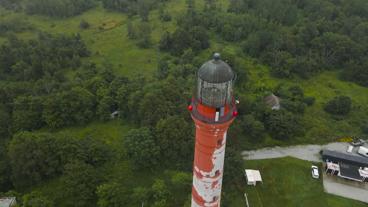 Aerial drone video revealing a old historic red colored lighthouse in Pakri towering over treetops and nature during a misty and foggy day at a summer peninsula. Renewable energy wind turbines at back